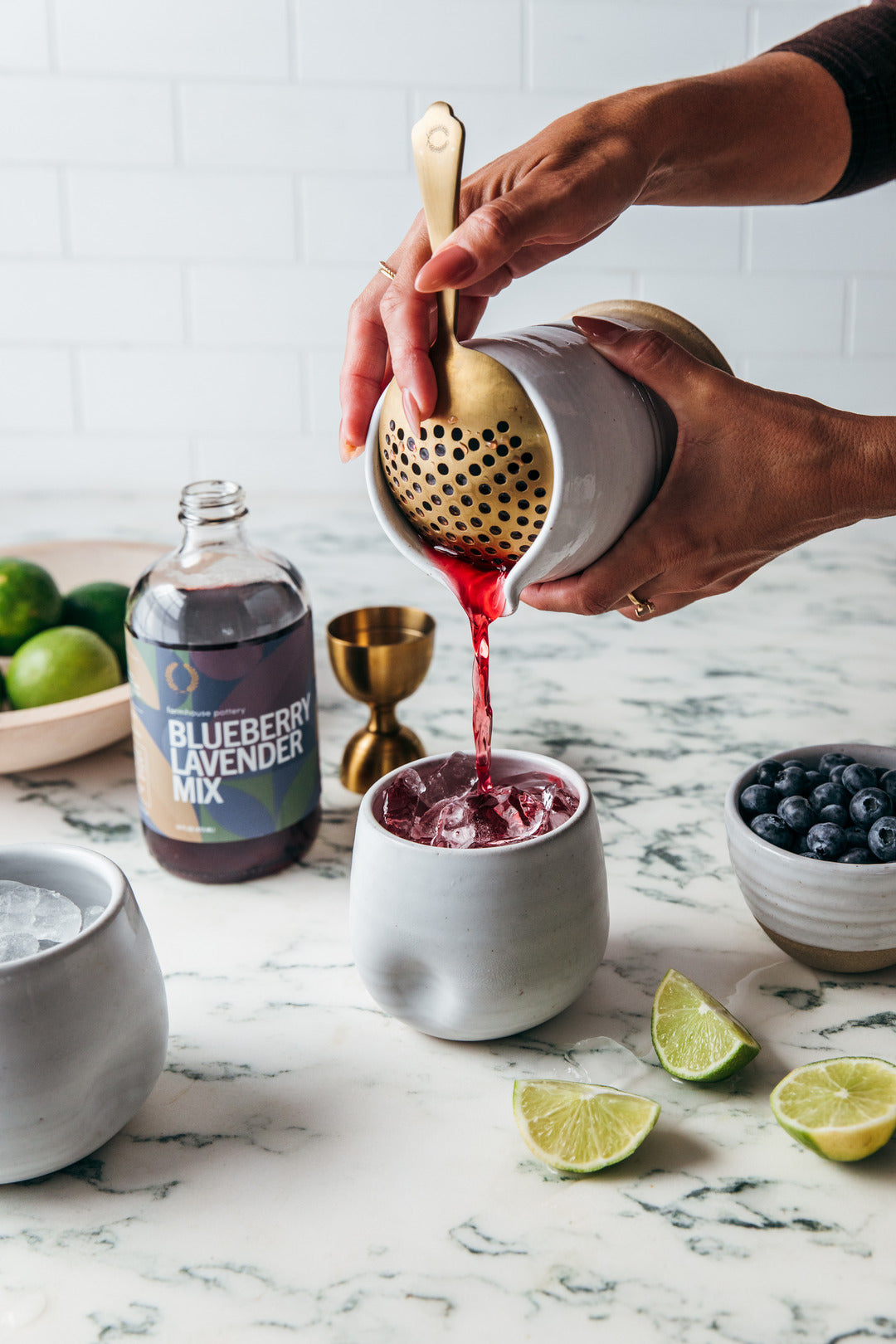 Hands pressing a red berry cocktail through the brushed-gold perforated strainer into a white ceramic cup, surrounded by limes and blueberries.