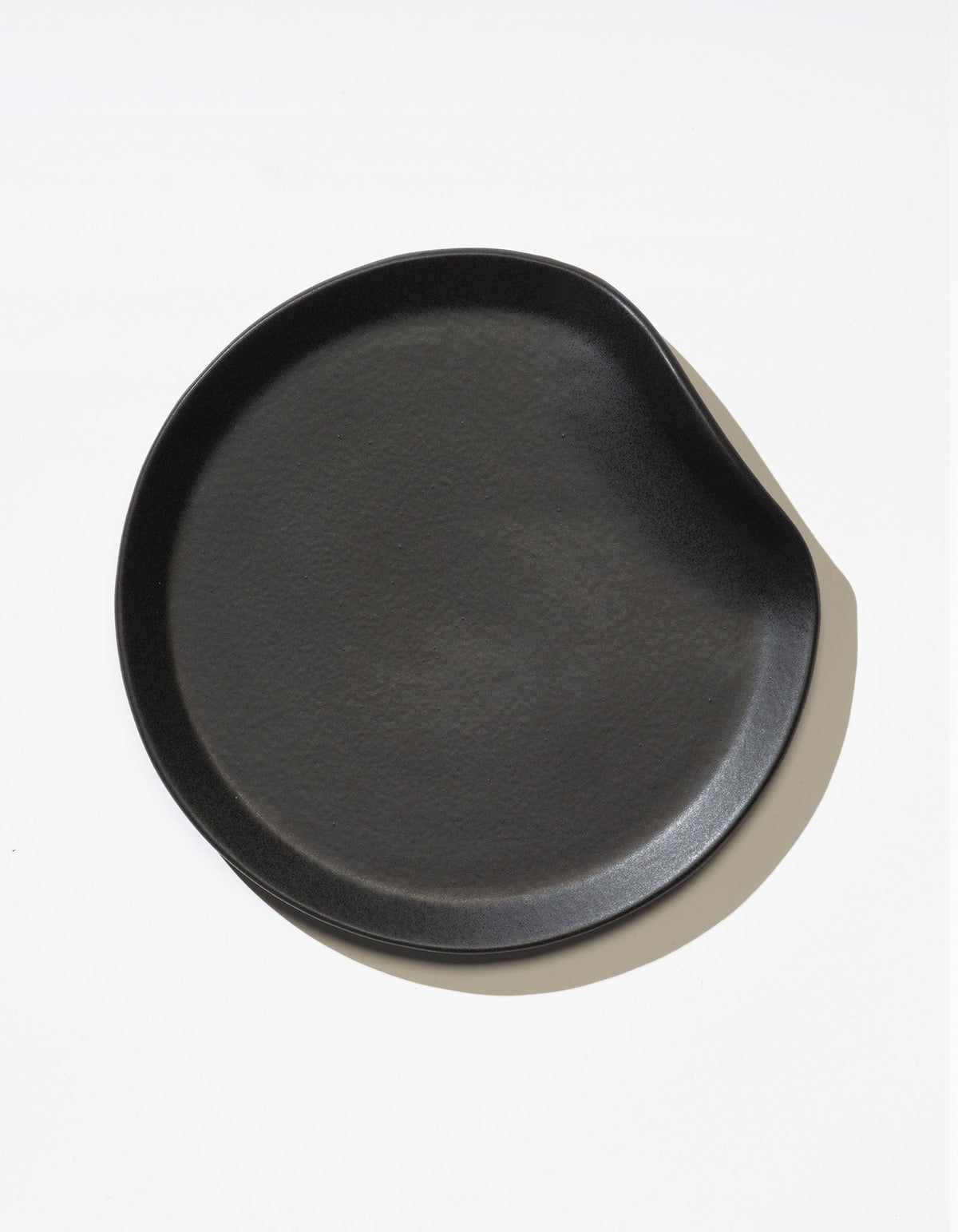 Tortuga Forma Salad Plate Single in Soot viewed overhead on white, showing a shallow serving surface, subtle texture and an inward curve along one side of the rim.