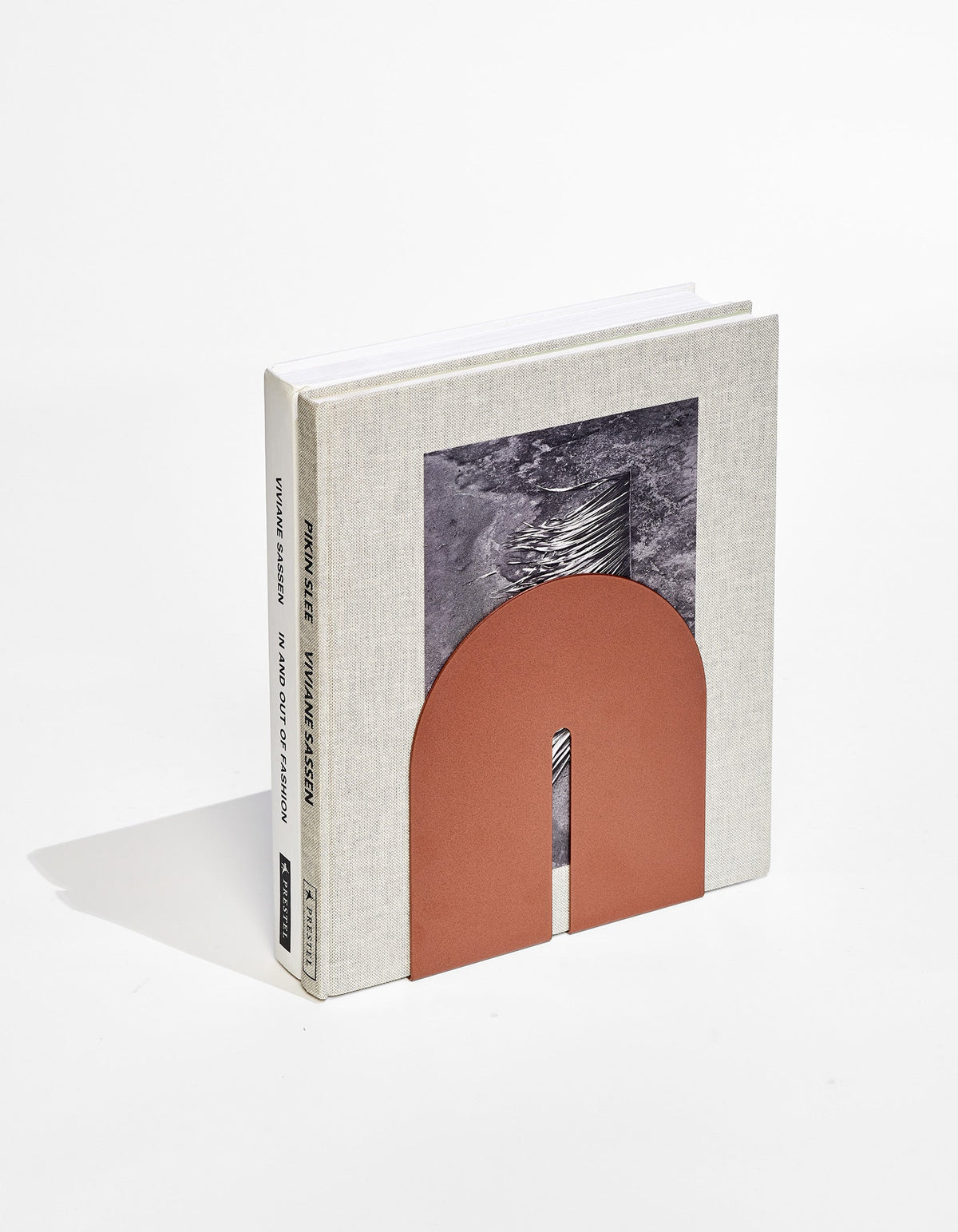Copper-coloured bookend from Tortuga Forma Dumbo Bookend Pairs - The Architect Collection supporting upright books on white, with its slotted arch resting against a grey cover with a black-and-white photograph.