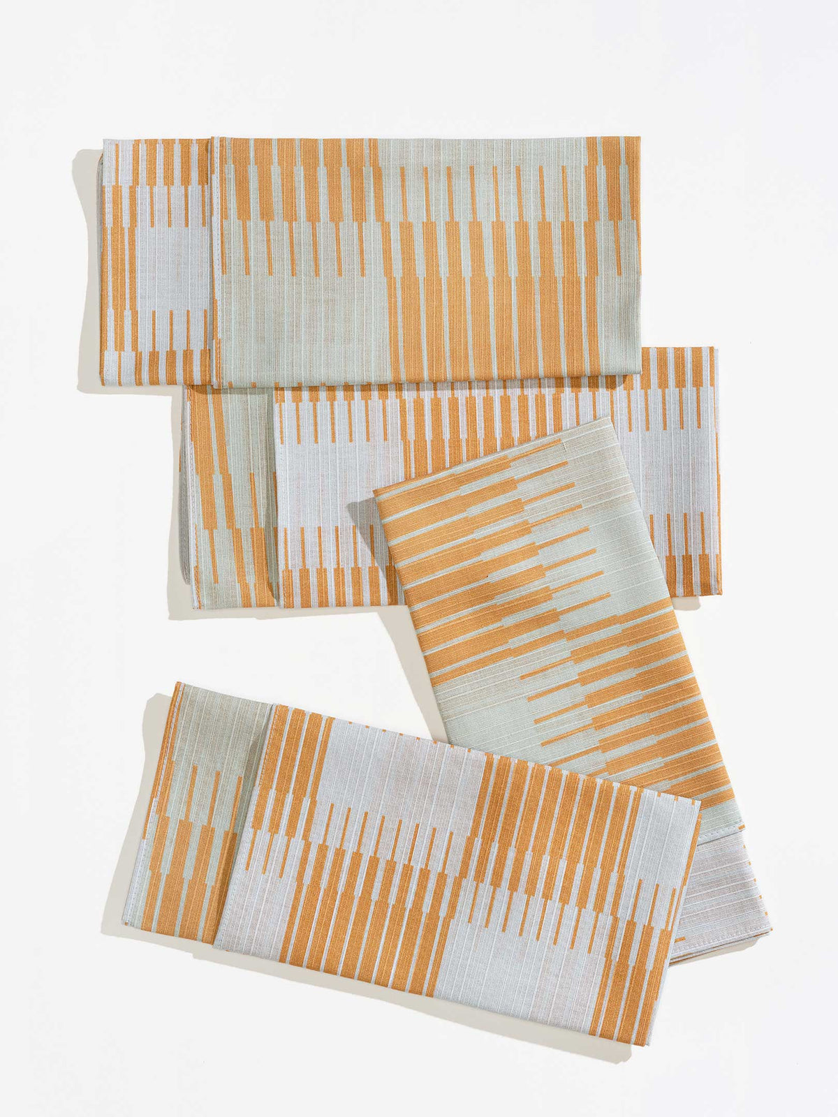 Four folded Tortuga Forma Dinner Napkins Double Sided Drift - Quiet Season arranged on white, showing orange and pale grey linear patterns forming staggered bands across the fabric.