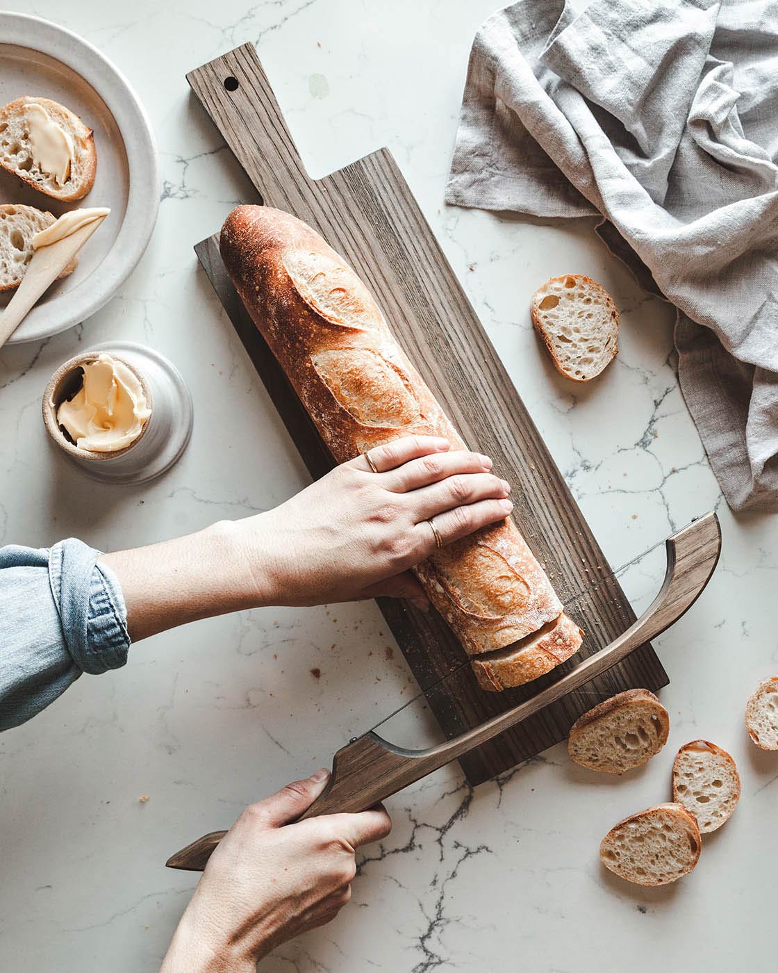 Overhead view of hands slicing a crusty baguette on a long dark wood Pantry Cutting Board beside butter, small bread slices, and a folded linen cloth.