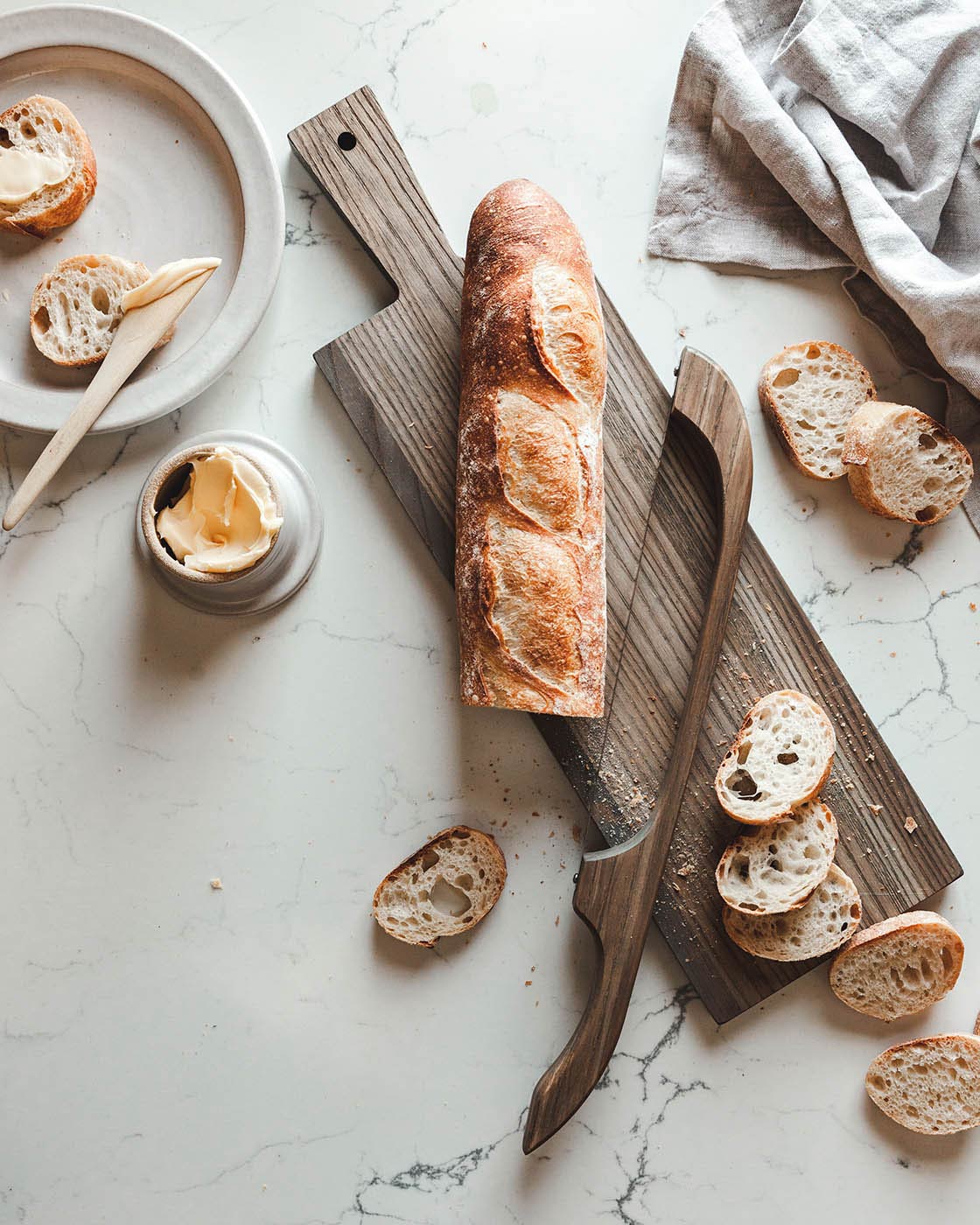 Crusty baguette and sliced rounds arranged with a dark wood bread bow on a long wooden cutting board.