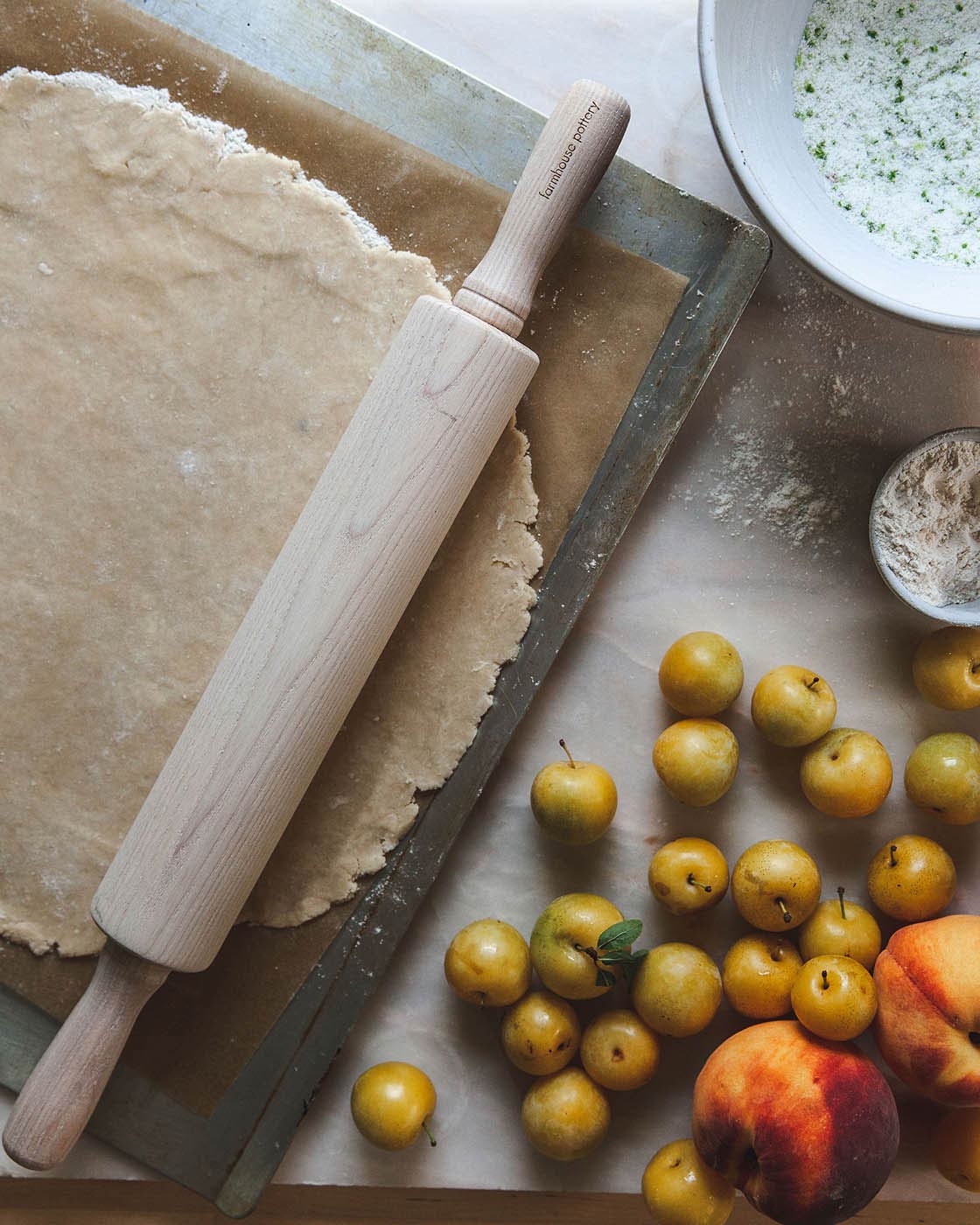 Gray-washed ash rolling pin resting beside rolled pastry dough on a baking sheet with small yellow plums and a red peach.