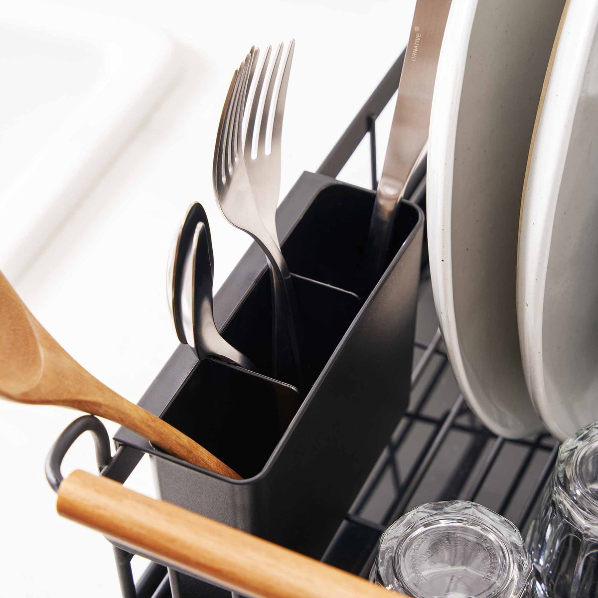 Close-up of the Yamazaki Home Dish Rack - Steel + Wood utensil caddy, showing separate black compartments holding a wooden spoon, cutlery and dark utensils beside upright plates and a wooden handle.