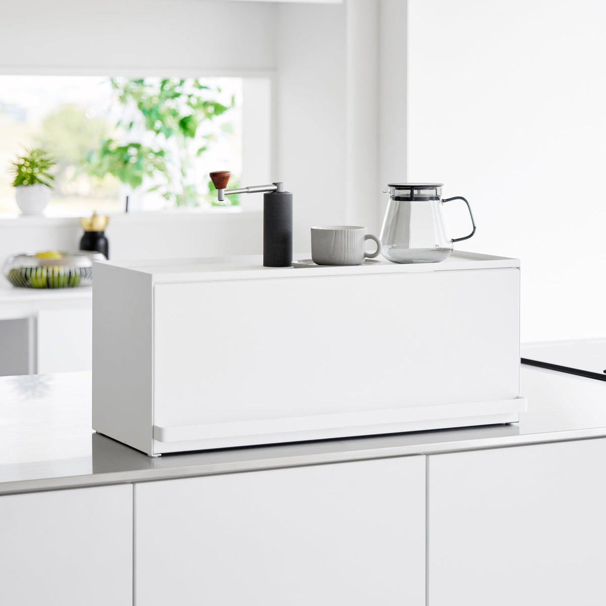 White Yamazaki Home Open-Back Countertop Storage Rack - Steel with its front panel closed on a bright kitchen counter, topped with a black coffee grinder, grey mug and glass carafe.