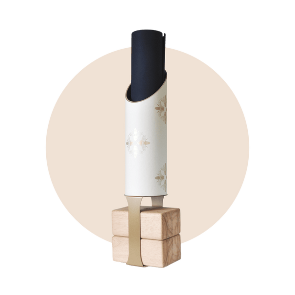 White cylindrical yoga storage tube decorated with delicate gold bee illustrations, holding a rolled black mat on four natural wood blocks with a tan support strap.