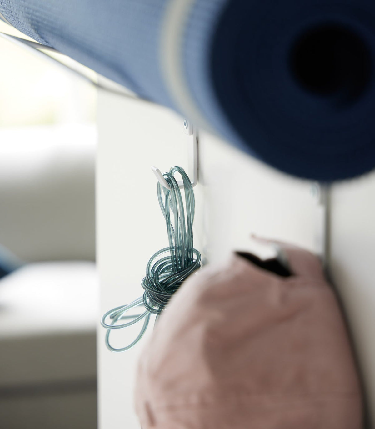 Detail of the white Yamazaki Home Over-the-Door Fitness Equipment Hanger - Steel lower hooks, with a turquoise jump rope and pink cap suspended beneath a rolled blue mat.