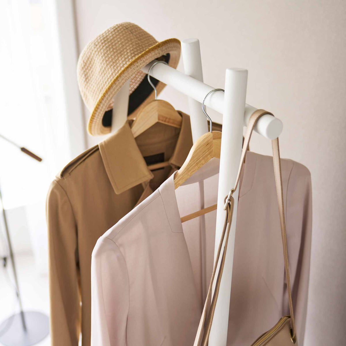 Close-up of the white Yamazaki Home Tri-Post Coat Rack (60" H) - Steel upper rail, showing cylindrical posts, projecting rail ends, wooden hangers, a straw hat and a shoulder-bag strap.
