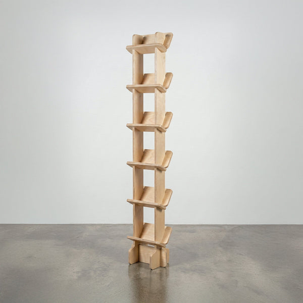 Studio Furnis Yankel tall narrow bookcase in natural wood with a central vertical spine and six pairs of sculptural angled shelves on a concrete floor