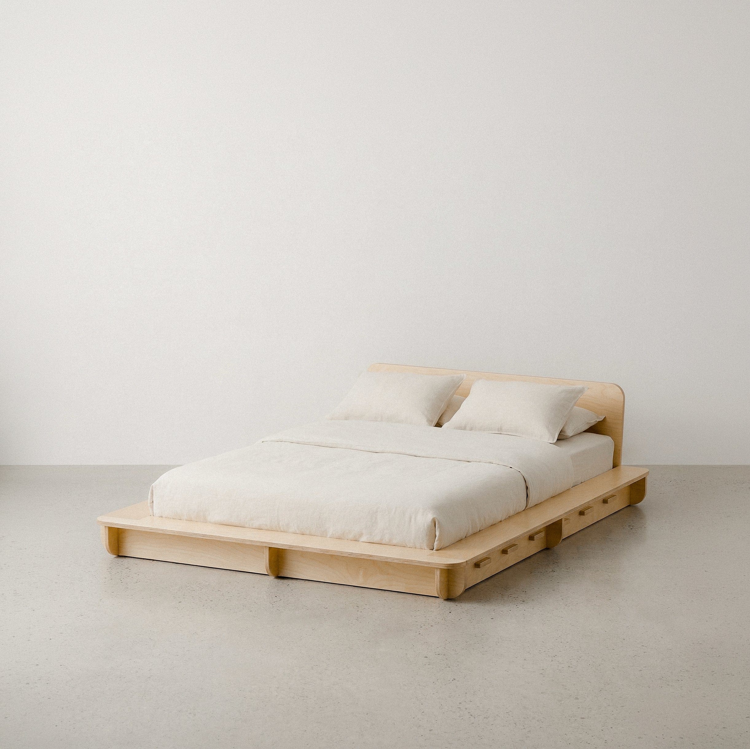 Studio Furnis natural wood bed platform with cream bedding and a low curved headboard, shown from the front-left against a studio background