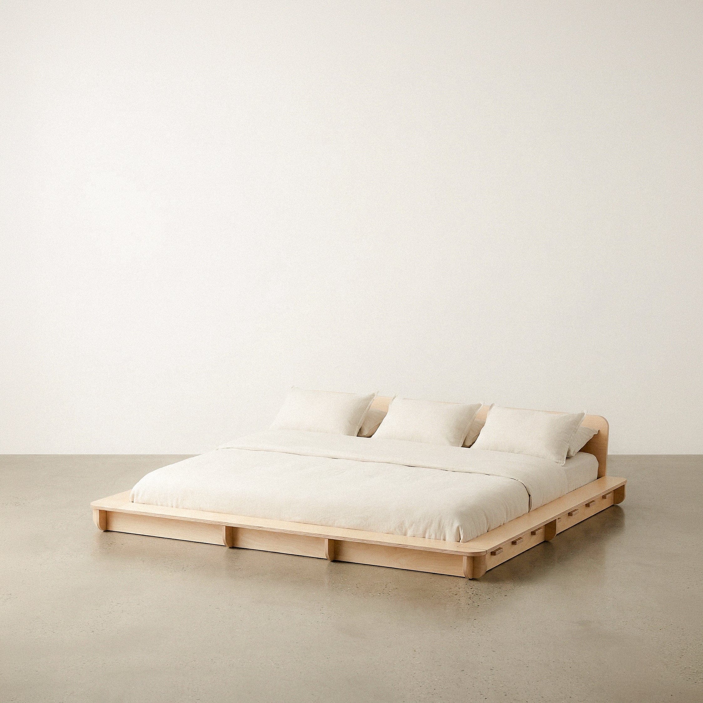 Extra-wide Studio Furnis natural wood bed platform with cream bedding, two pillows and a low curved headboard against a neutral studio background