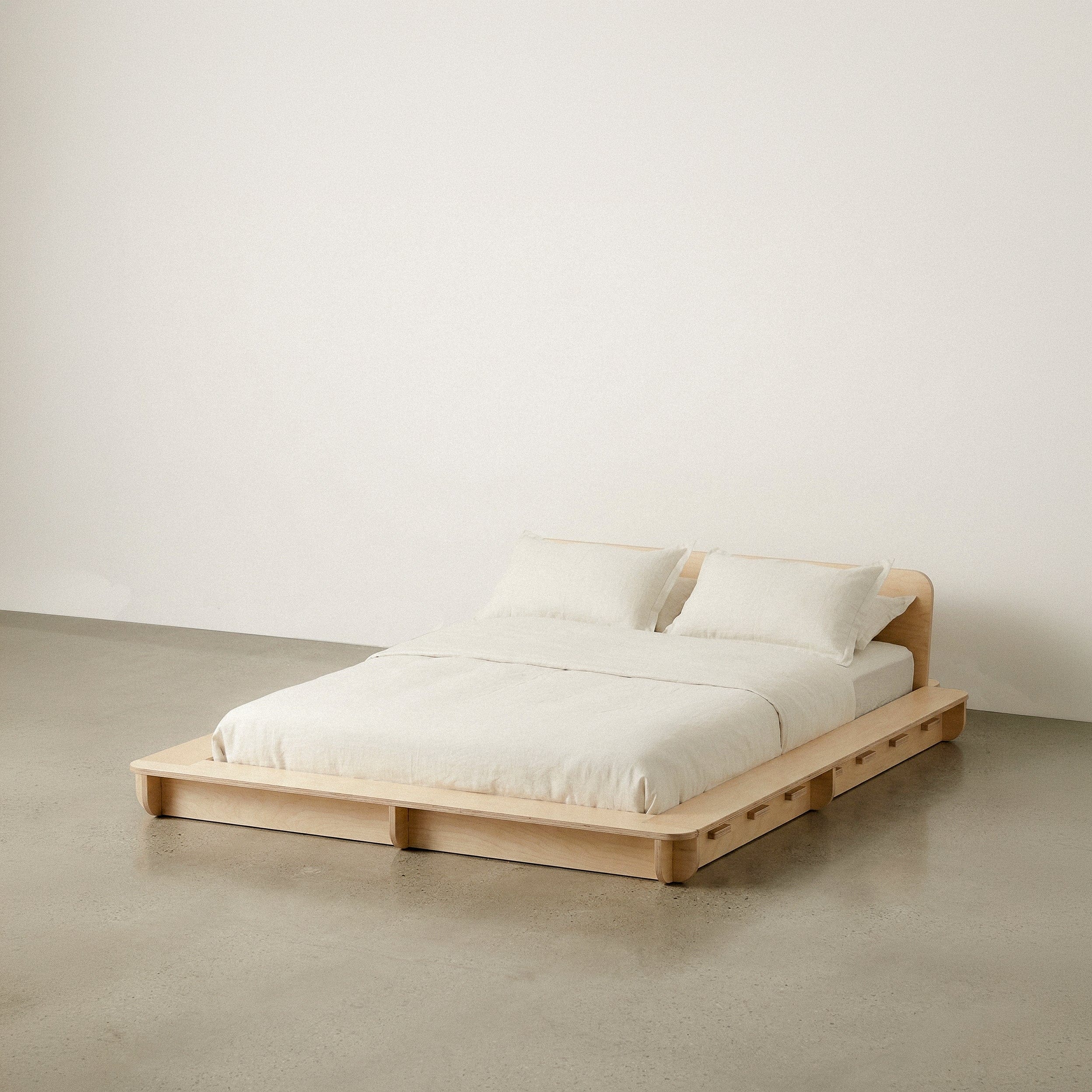 Wide Studio Furnis wood bed platform with cream bedding, one pillow and a low curved headboard against a neutral studio background