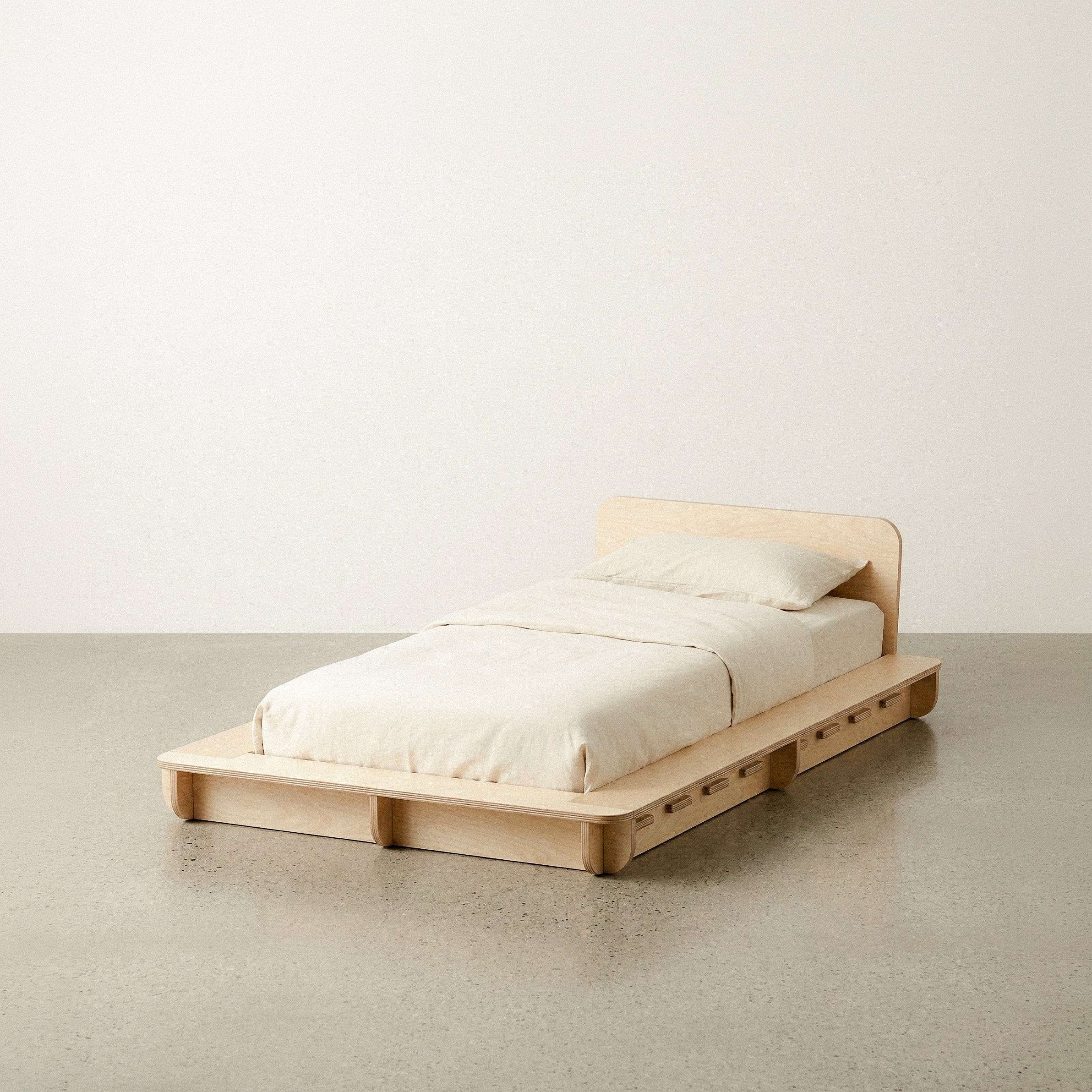 Narrow Studio Furnis natural wood bed platform with cream bedding and a curved wood headboard against a plain studio background