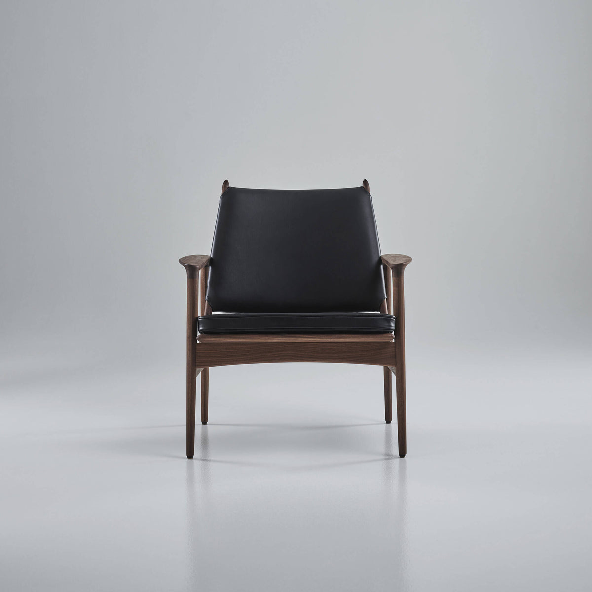 Front view of the black leather Eikund Broadway Lounge Chair with a brown wood frame in a gray studio, showing rounded armrest ends and slender legs.