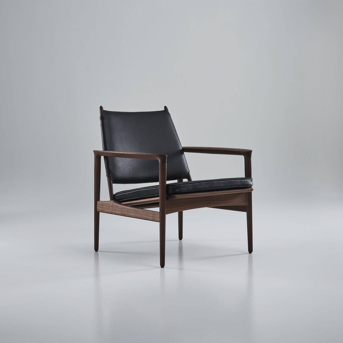 Angled front view of the black leather Eikund Broadway Lounge Chair in a gray studio, showing its open sides, gently reclined backrest, and thin seat cushion above the wood rails.