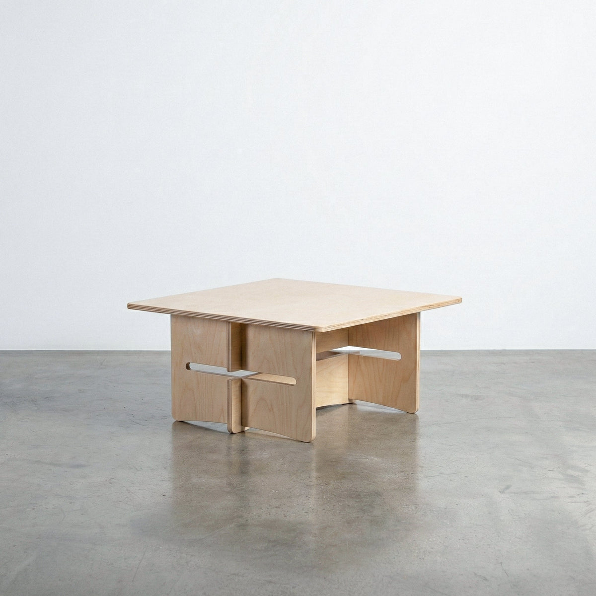 Williamsburg natural wood coffee table with a broad square top and sculptural interlocking panel base with projecting horizontal supports on a concrete floor