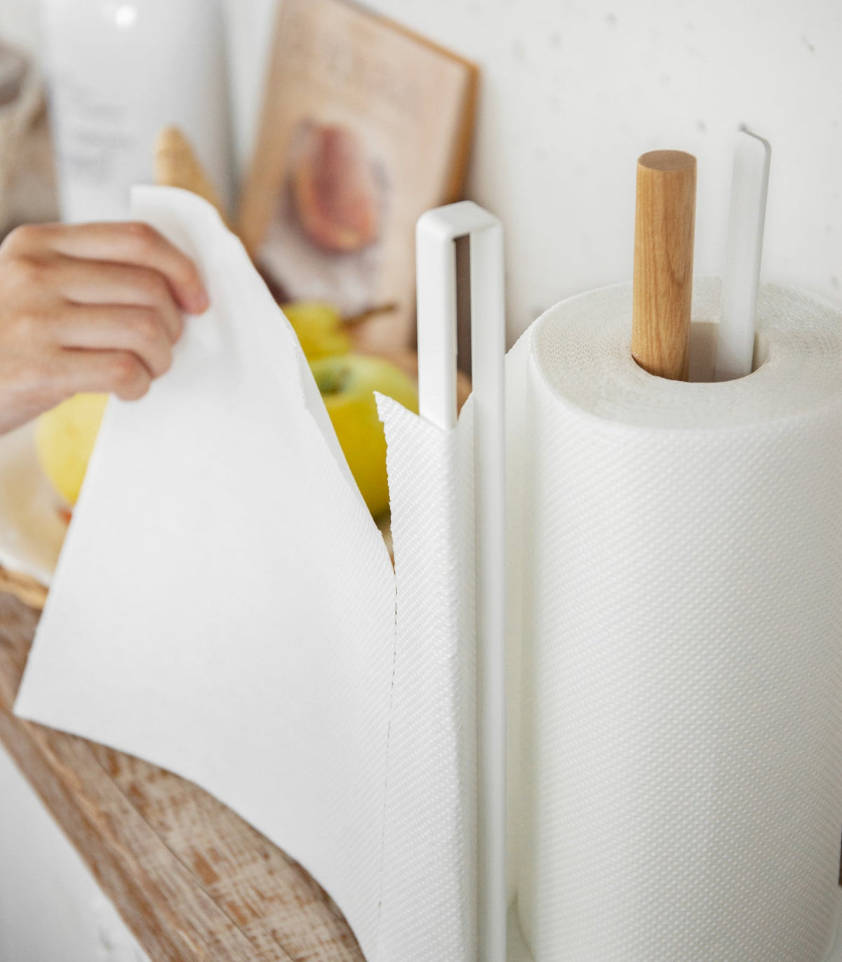 Close-up of a person pulling a paper towel sheet leftward from the Yamazaki Home Paper Towel Holder - Steel + Wood, with the roll resting beside the tall white side rail.
