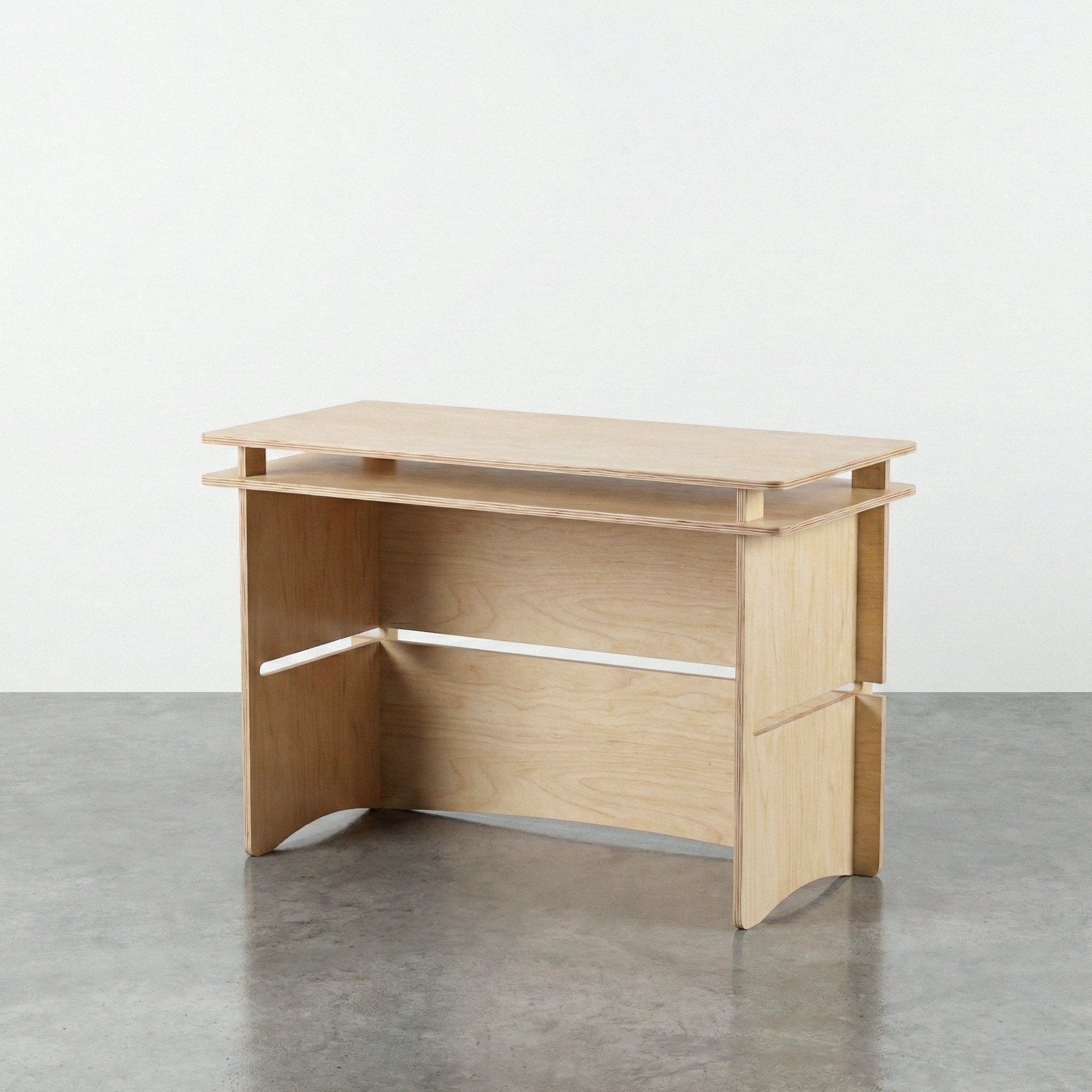 Studio Furnis Williamsburg natural wood desk with a raised desktop shelf, broad panel legs, open knee space and a rear modesty panel on a concrete floor