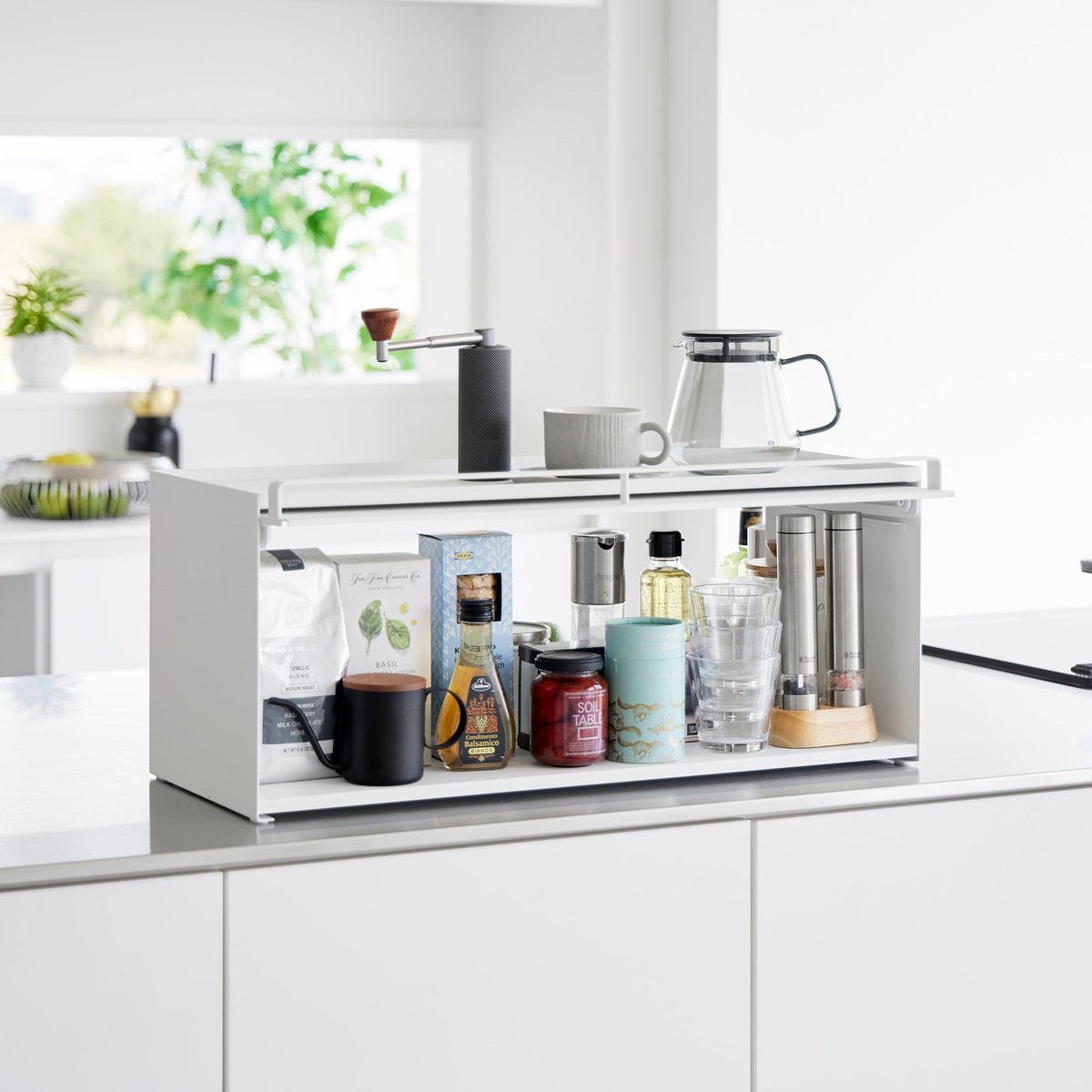 White Yamazaki Home Open-Back Countertop Storage Rack - Steel on a kitchen counter with its front panel raised, revealing bottles, jars, drinkware and tall mills; a coffee grinder, mug and glass carafe sit on top.