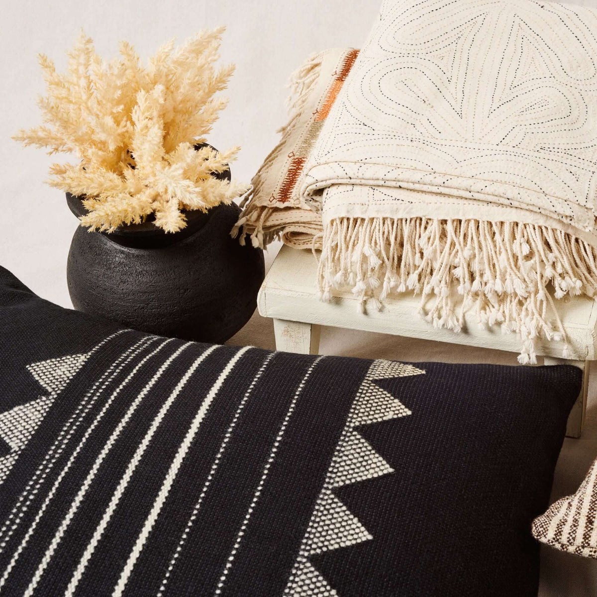 Kora Black pillow styled with a black vase of dried grasses and layered cream fringed throws
