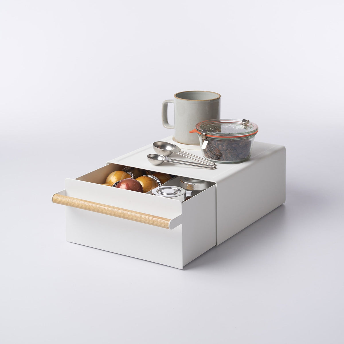 White Yamazaki Home Countertop Rack with Drawer - Steel + Wood with its wood-handled drawer partly open, holding coffee capsules and small containers; a mug, glass jar and measuring spoons rest on top.