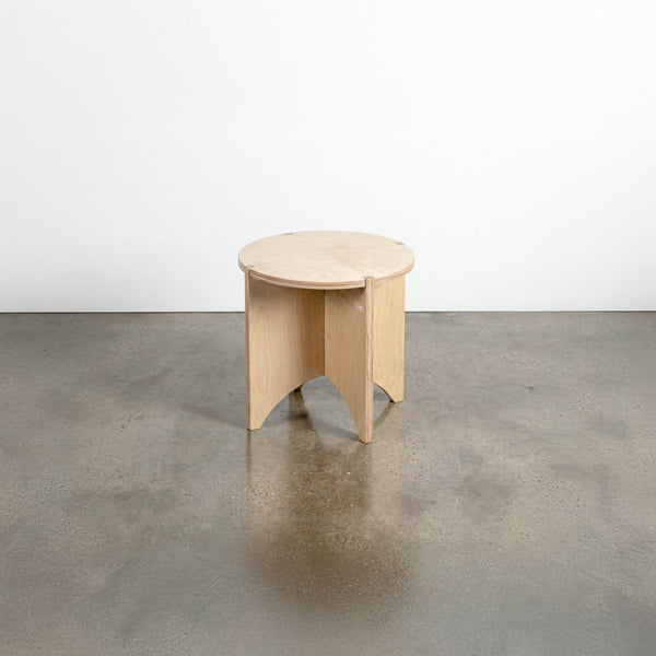Studio Furnis YOYO natural wood end table with a round top and sculptural intersecting panel legs, displayed on a polished concrete floor