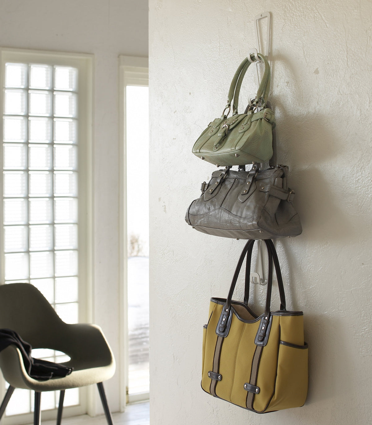 White Yamazaki Home Cascading Storage Hooks - Steel mounted against a pale wall, holding green and grey handbags above a yellow tote near a doorway and black chair.
