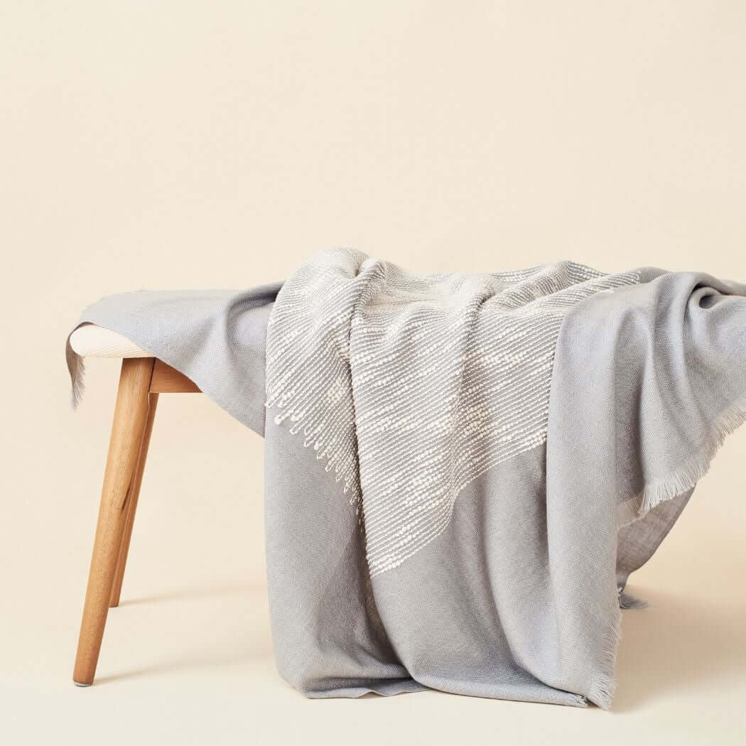 Variously Flo Gray merino throw draped over a pale wood bench, showing its soft gray field, white textured pattern and fringed edges