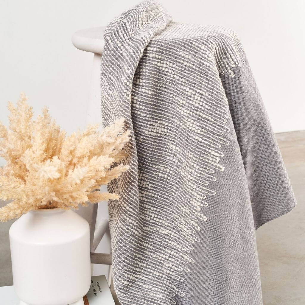 Flo Gray handloom throw folded over a chair beside a white vase of dried grasses, highlighting its woven white detailing