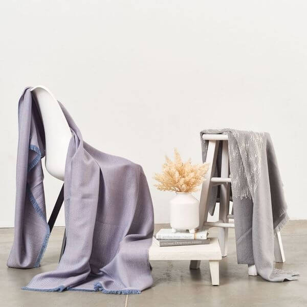 Flo Gray throw displayed with coordinating lavender and gray textiles draped over chairs in a minimalist setting