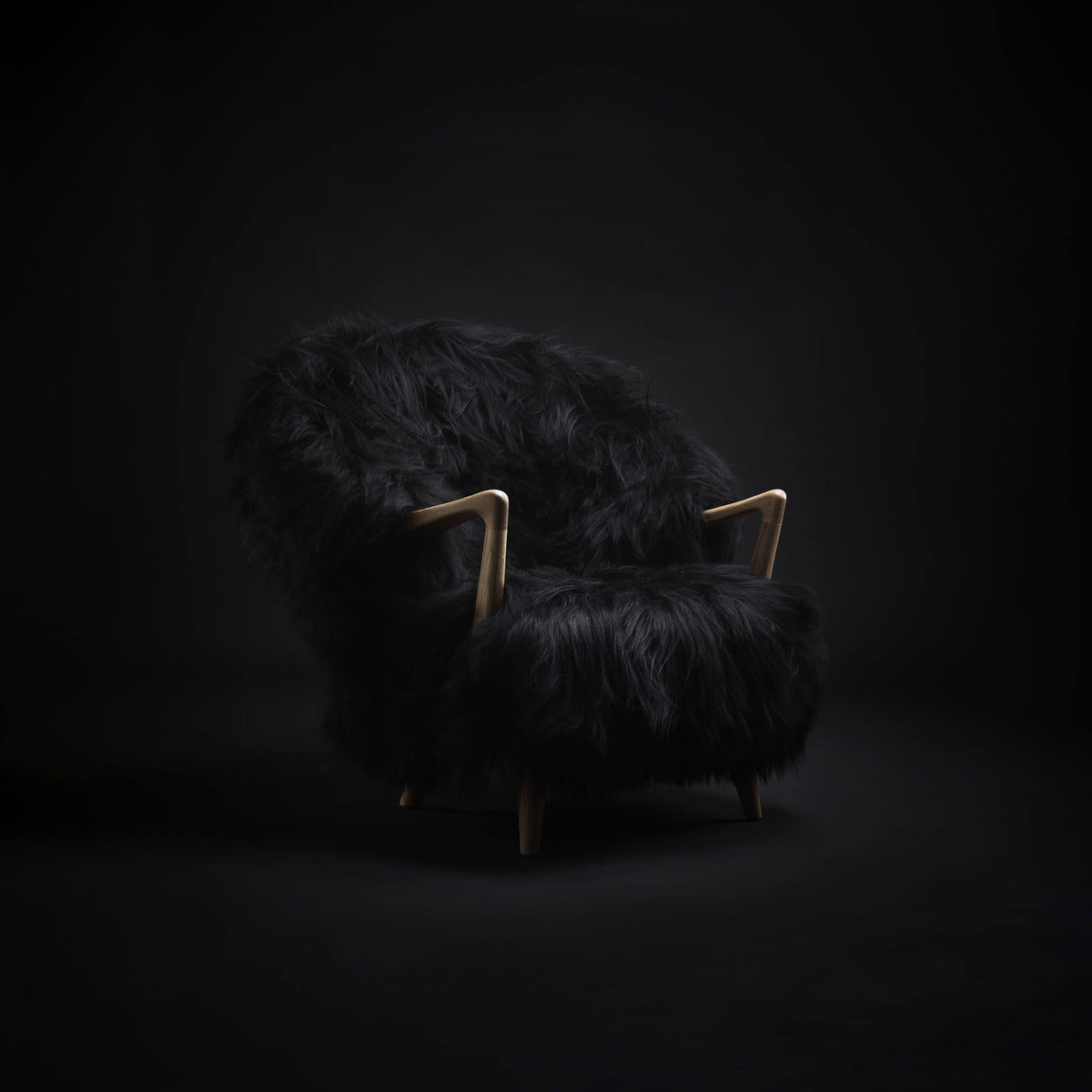 Eikund Fluffy Lounge Chair in a dim black studio, with directional light picking out the long black upholstery and curved light wood armrests.