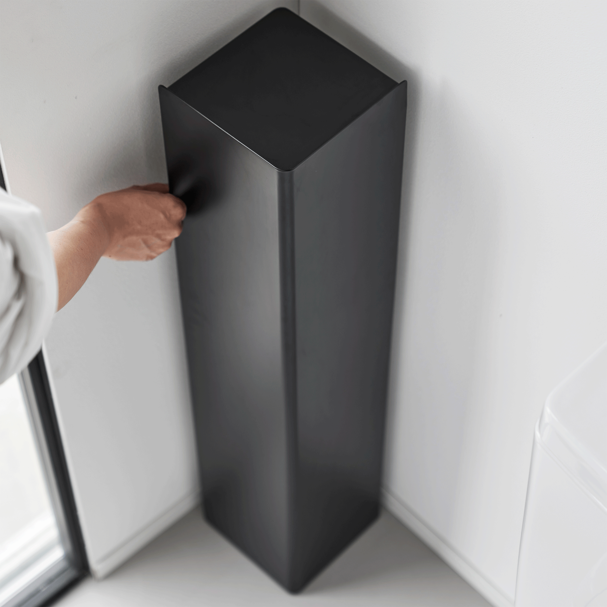 A person turns the empty black Yamazaki Home Rolling Bathroom Organizer - Steel in a bathroom corner, showing its tall rectangular panels and square top from above.