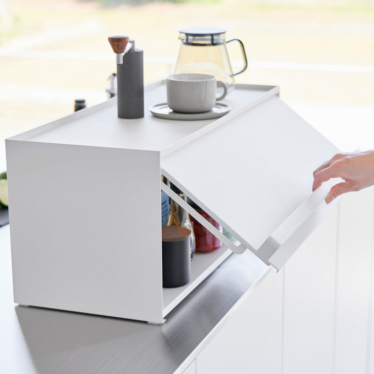 Side view of the Yamazaki Home Open-Back Countertop Storage Rack - Steel as a person holds its front panel tilted outward, revealing the storage compartment beneath the fixed top.