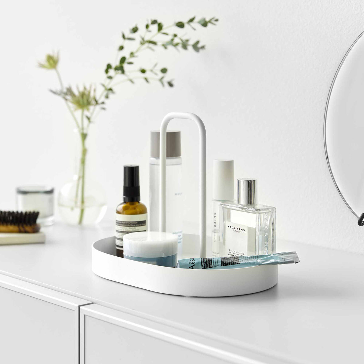 White Yamazaki Home Vanity Tray with Handle - Steel organizing skincare bottles, jars, a clear perfume bottle and a toothbrush on a bathroom counter beside a mirror.