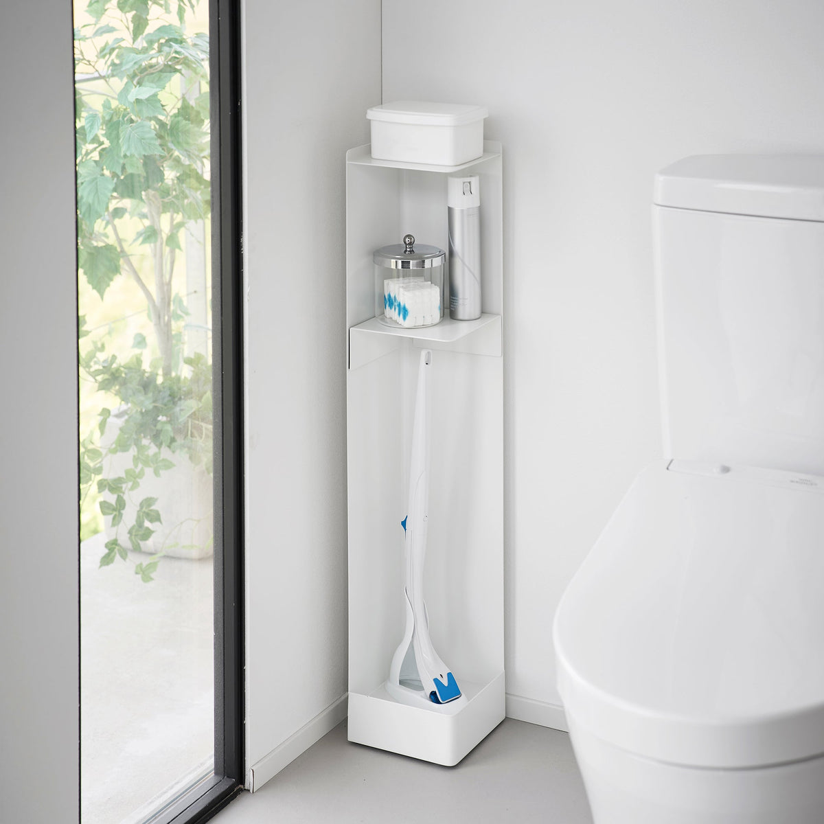 White Yamazaki Home Rolling Bathroom Organizer - Steel beside a toilet and window, with a tissue box on top, small bathroom supplies on the upper shelf and long cleaning tools in the lower compartment.