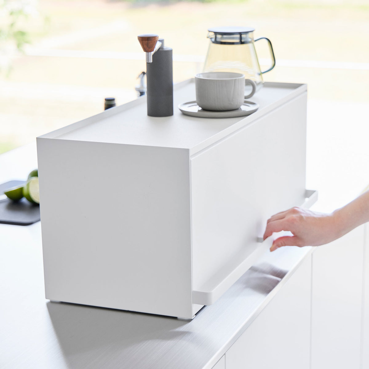 Close-up of a hand gripping the lower front handle of the closed Yamazaki Home Open-Back Countertop Storage Rack - Steel, with coffee-making items arranged on top.