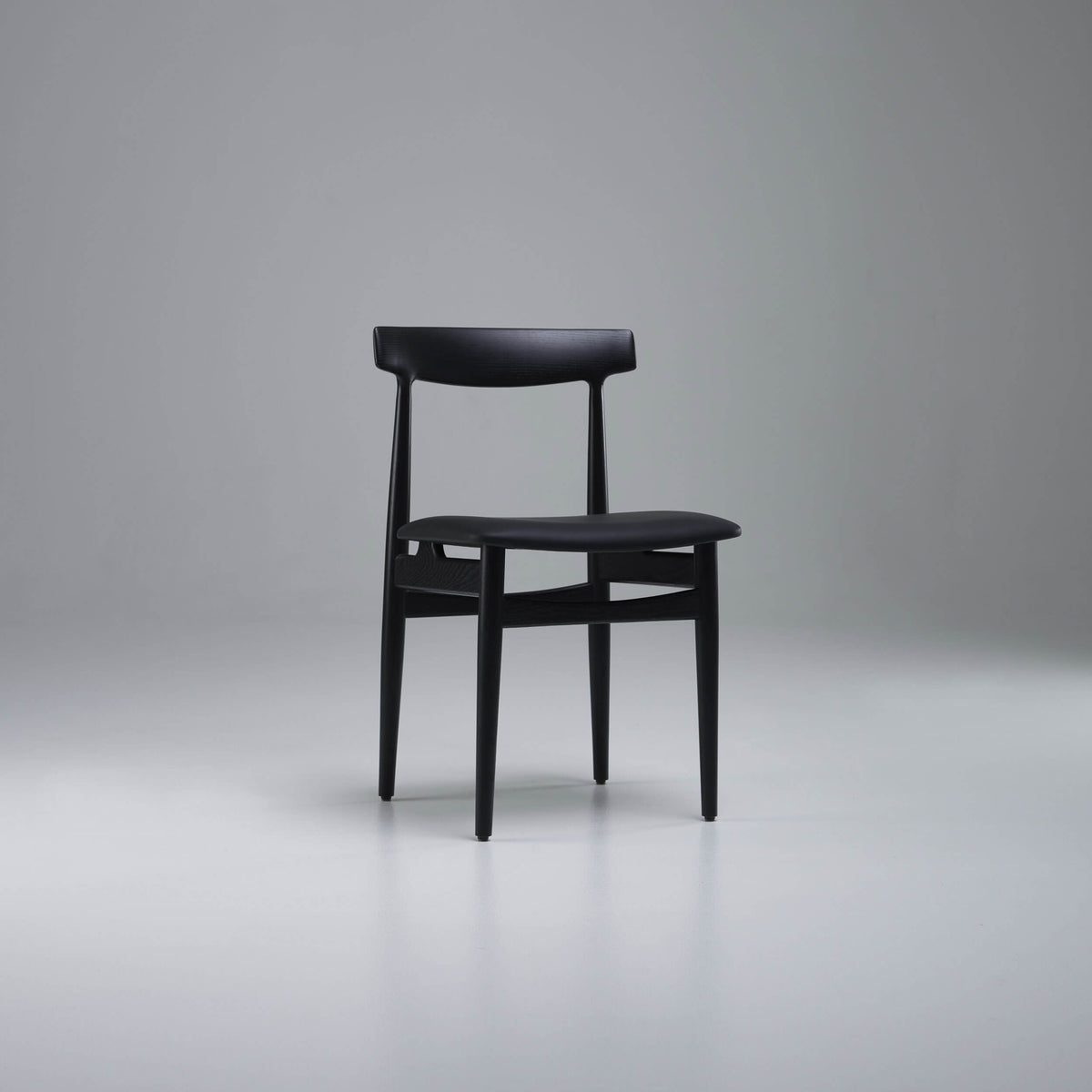 Three-quarter front view of the all-black Eikund Hertug Dining Chair in a gray studio, showing its curved backrest, slim padded seat, and exposed wood frame.