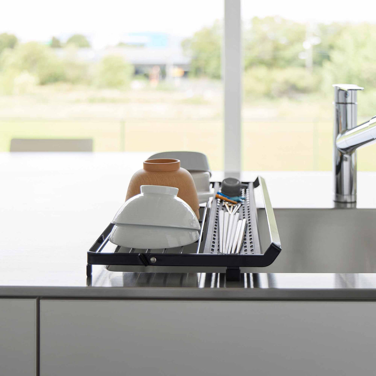 End view of the black Yamazaki Home Slim Over-the-Sink Dish Drying Rack - Steel over a sink, holding pale bowls and brown cups beside the narrow utensil tray.