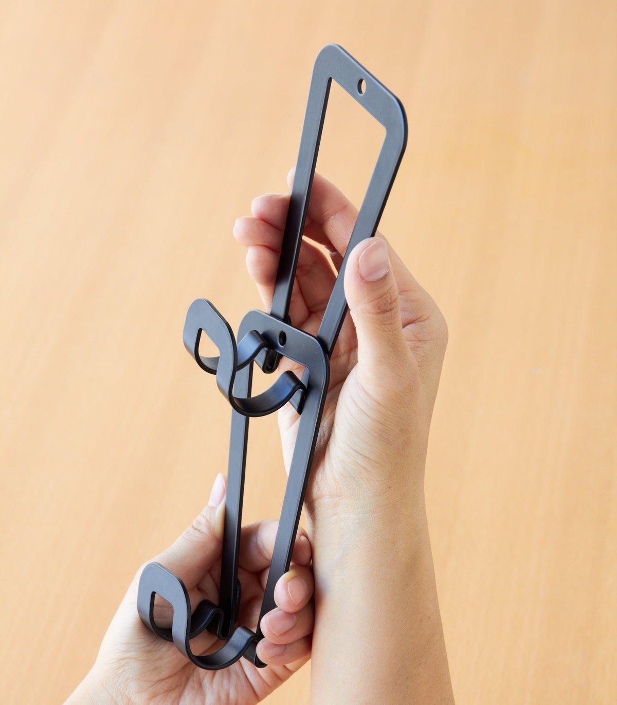 Close-up of two hands holding black Yamazaki Home Cascading Storage Hooks - Steel, showing how the elongated frames link together beside the projecting curved hooks.