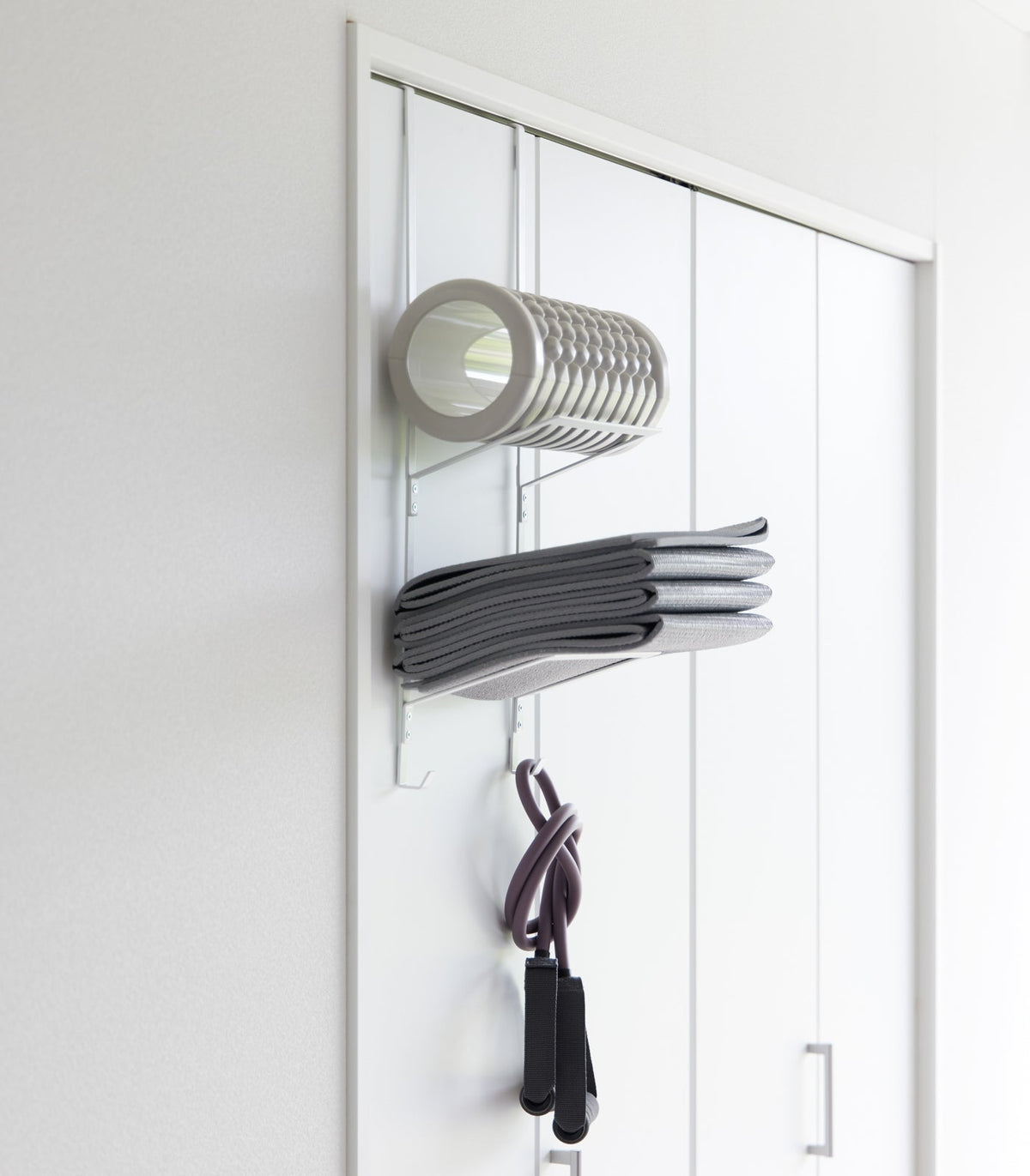 White Yamazaki Home Over-the-Door Fitness Equipment Hanger - Steel mounted on a white sliding closet door, holding a foam roller, folded grey mat and purple resistance bands.