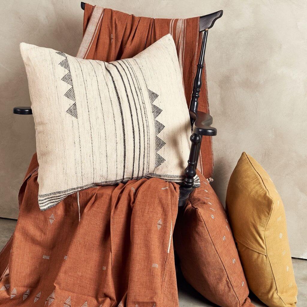 Kora pillow displayed on a rustic wooden chair draped with rust-colored fabric.