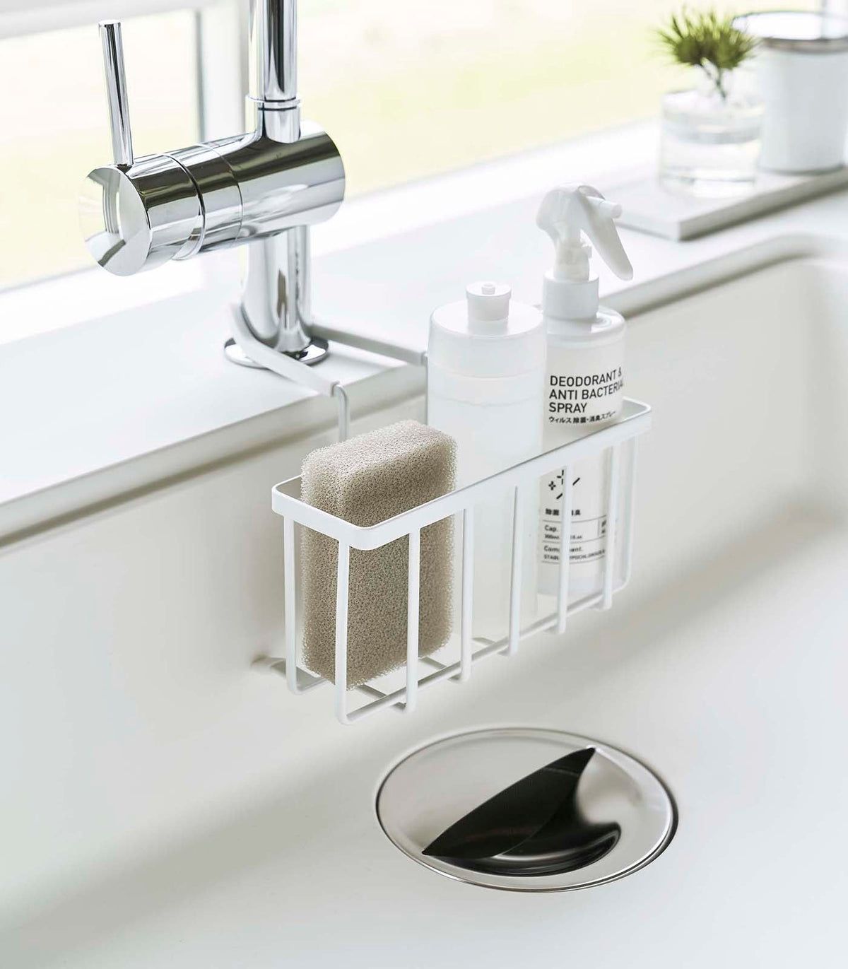 White Yamazaki Home Faucet-Hanging Sponge Caddy - Steel suspended inside a white sink beneath a chrome faucet, holding a beige sponge, white dispenser and spray bottle in its open wire basket.