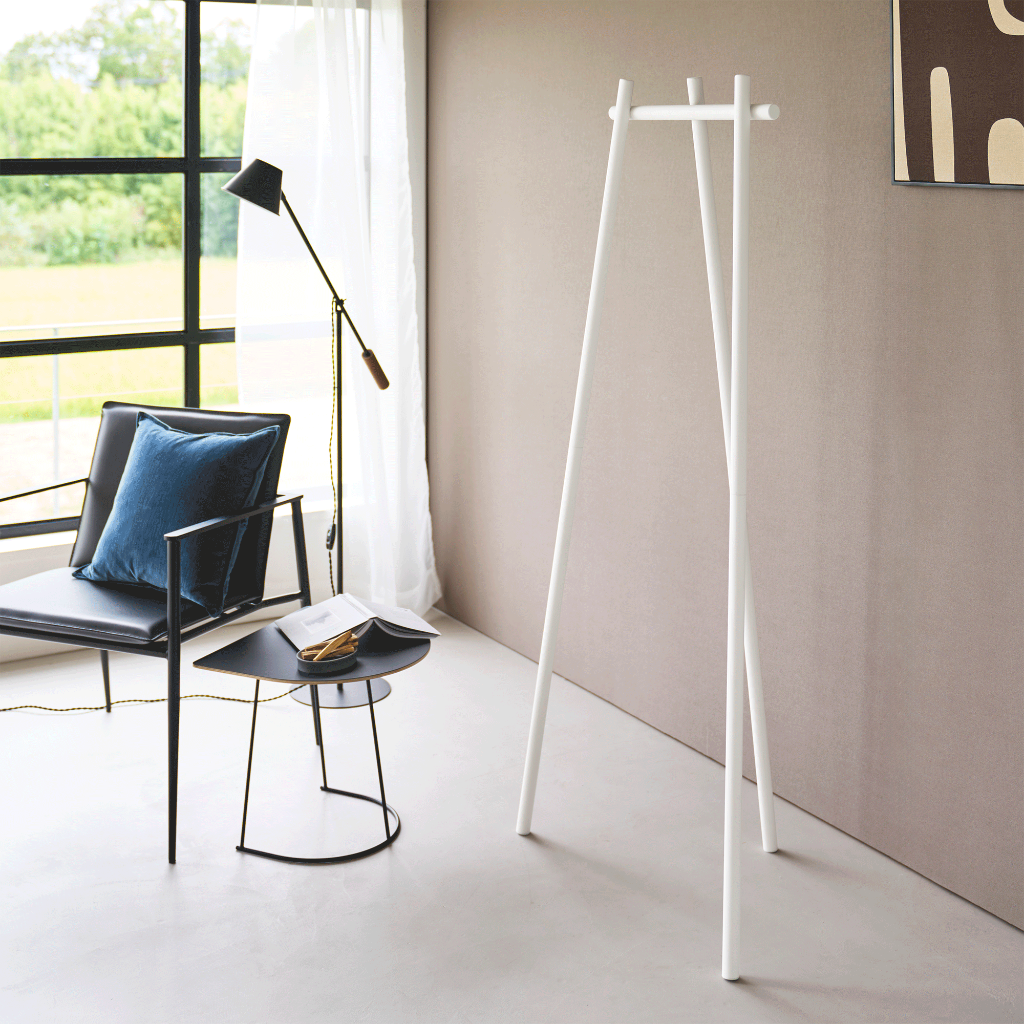 White Yamazaki Home Tri-Post Coat Rack (60" H) - Steel holding a tan trench coat and straw hat beside a beige wall, near a window, black chair with blue cushion and small side table.