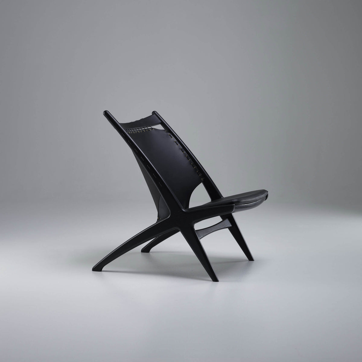 Three-quarter side view of the black Eikund Krysset Lounge Chair in a gray studio, showing its tall sloping back, padded seat, and splayed crossed supports.