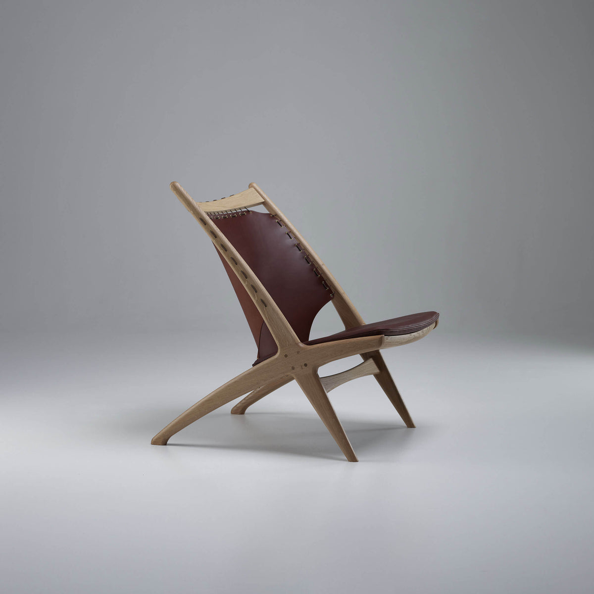 Side view of the Eikund Krysset Lounge Chair with a light wood frame and reddish-brown leather, showing the open triangular space beneath the reclined back and its X-shaped base.