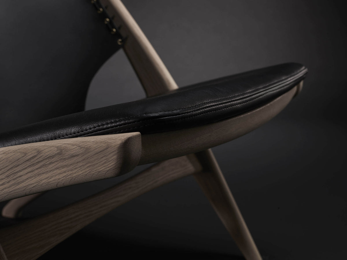 Close-up of the Eikund Krysset Lounge Chair’s black seat cushion, showing its stitched rounded edge above a curved wood seat rail and angled frame.