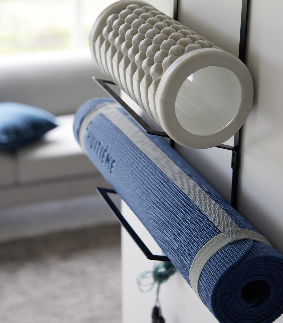 Angled close-up of the black Yamazaki Home Over-the-Door Fitness Equipment Hanger - Steel supporting a textured grey foam roller above a blue exercise mat secured with grey straps.