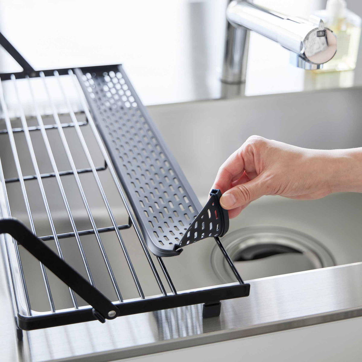 A hand lifts the black perforated utensil tray from the Yamazaki Home Slim Over-the-Sink Dish Drying Rack - Steel, showing the removable section above the sink.