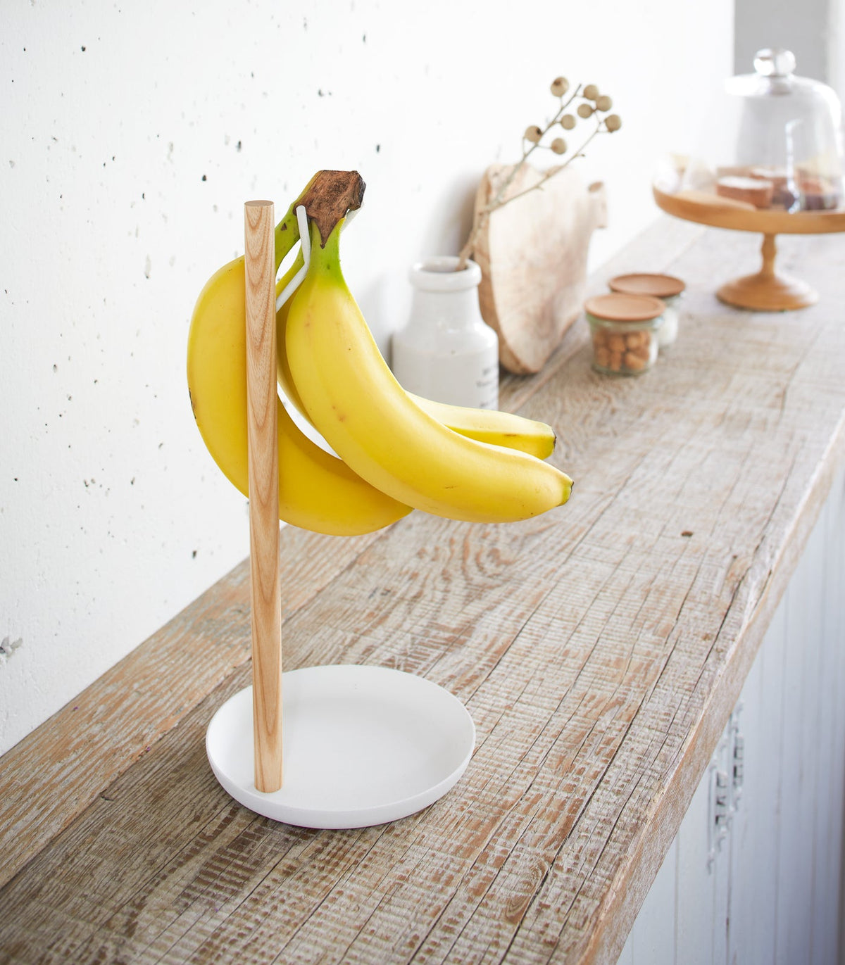 Angled view of the Yamazaki Home Banana Stand - Steel + Wood on a rustic wooden kitchen counter, with yellow bananas suspended above its white dish-shaped base.