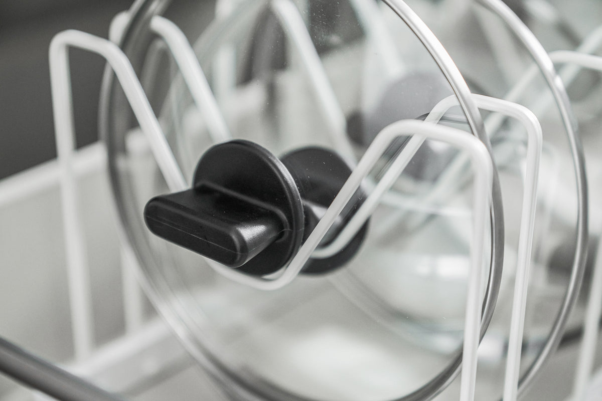 Detail of a glass lid in the white Yamazaki Home Adjustable Pot Lid Organizer - Steel, showing its black handle resting within a dip in the wire divider.